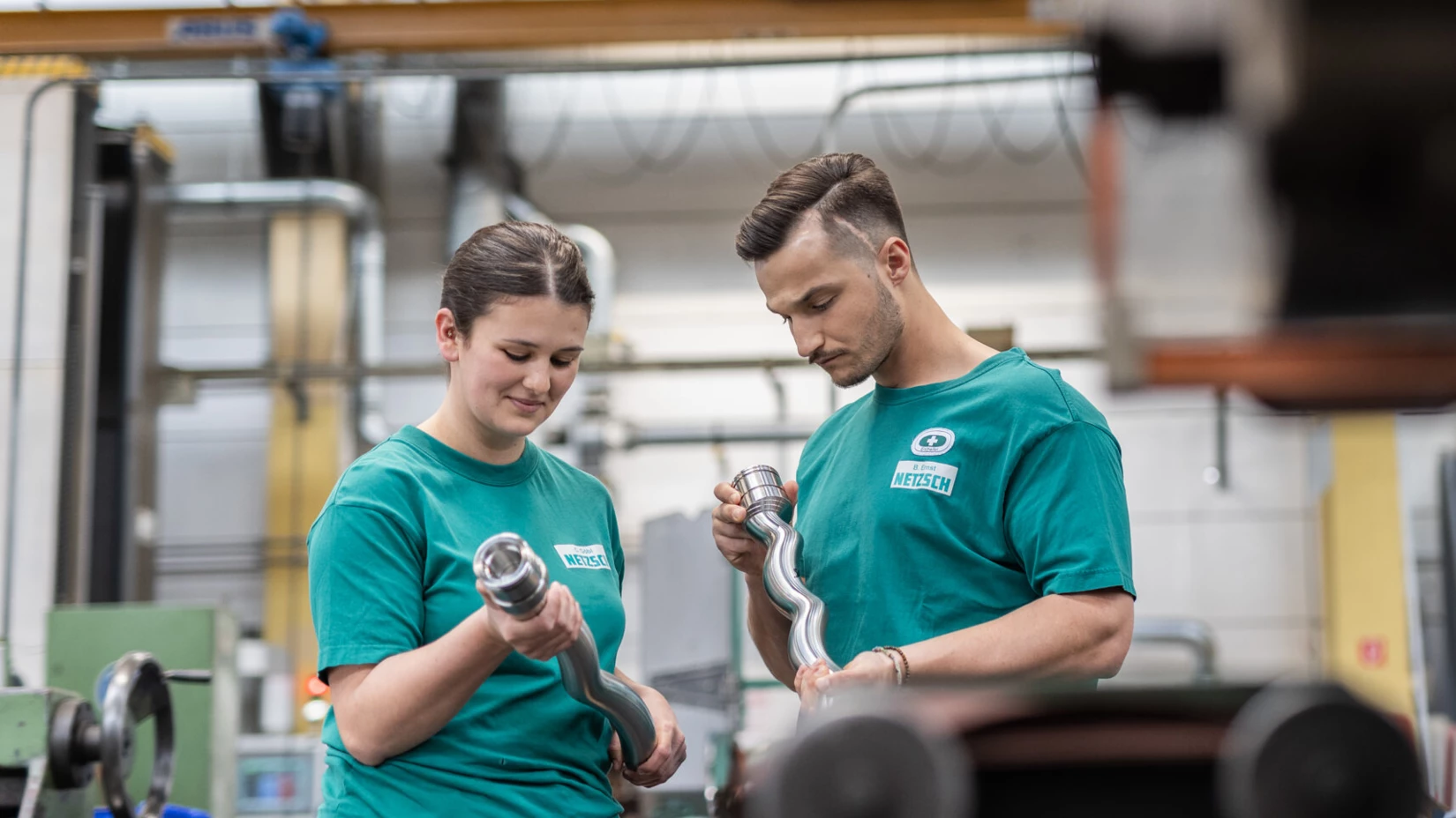 Two technicians in green shirts examine metal components in a manufacturing workshop, showcasing teamwork and precision.